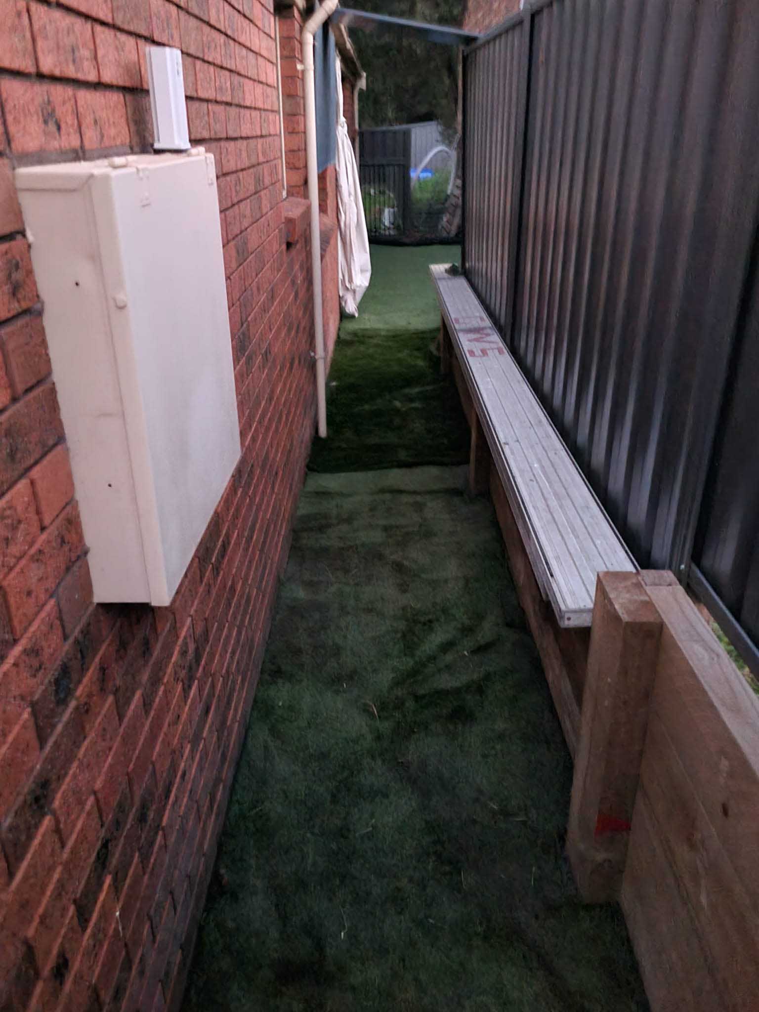 Narrow side passage beside a brick house with artificial turf and timber edging